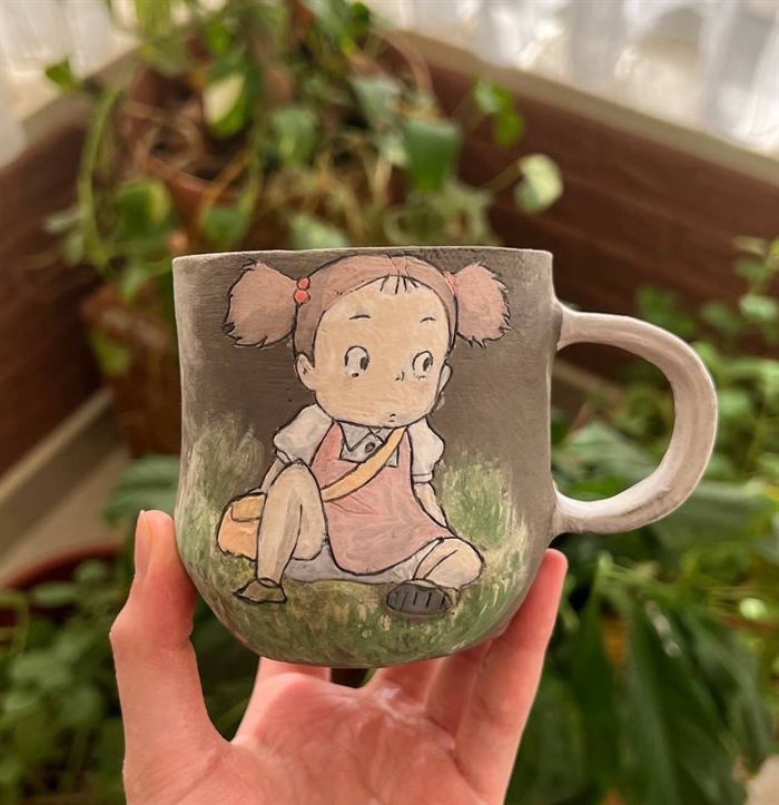 Grey stoneware mug carved and painted with a small girl in a pink pinafore sitting on green grass.