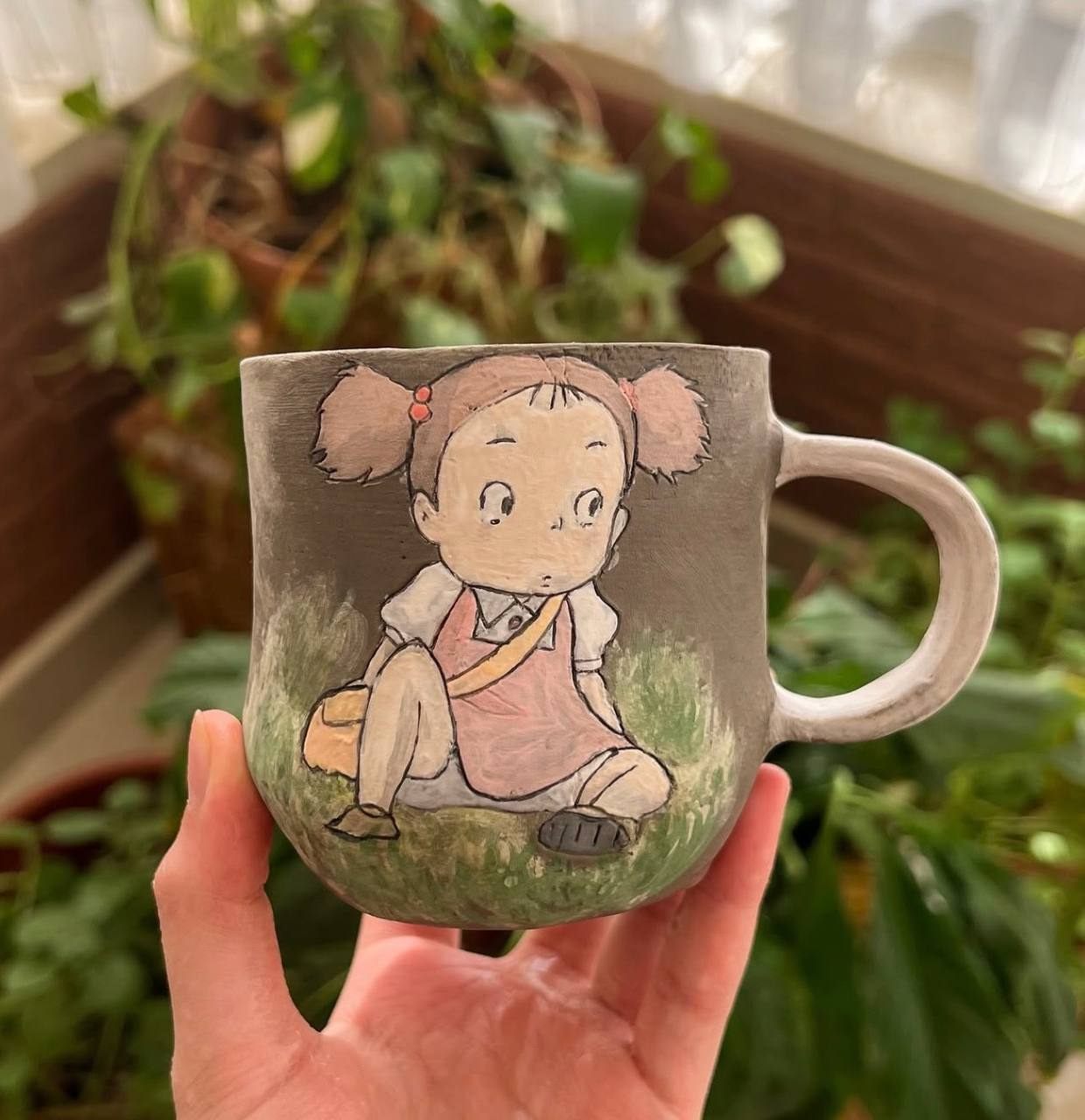 A hand-built stoneware mug held in the light, carved with a small girl in a pink pinafore sitting on grass.