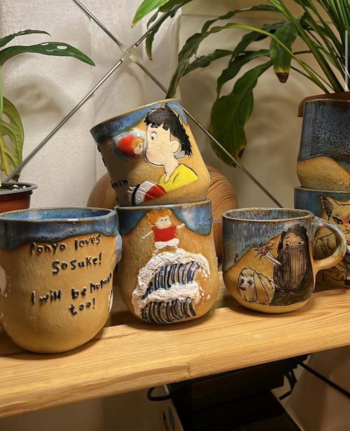 A wooden shelf holding several sandy-glazed cups with blue drip rims, each carved with a different painted character.