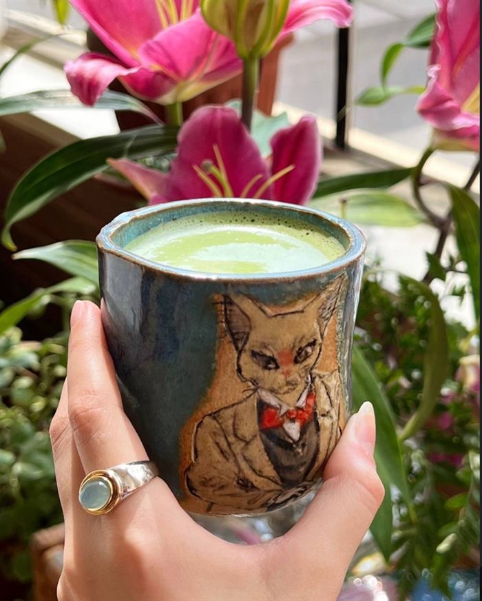 Hand holding the blue cat tumbler in warm light, pink lilies blooming behind it.